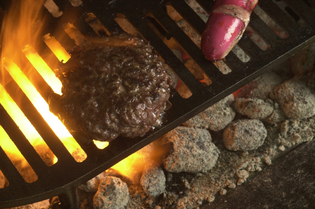 Hamburger and hot dog grilling over charcoal briquettes on a BBQ.