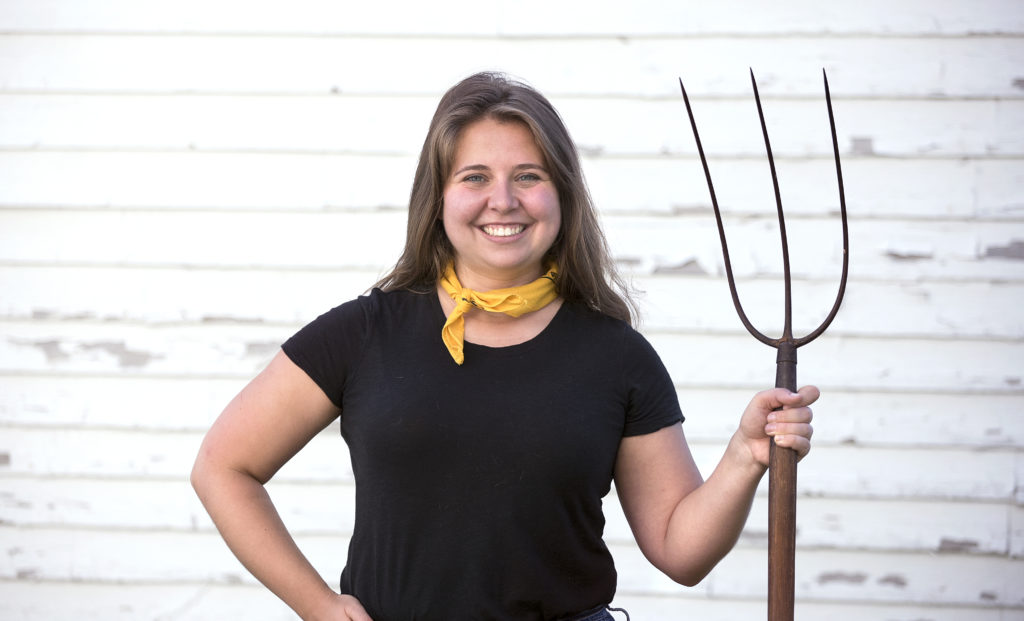 Sam Schipani is shown standing in front of a white, clapboard building holding a pitchfork. She is the writer behind Sam Tries Things, a new column on Hello Homestead.
