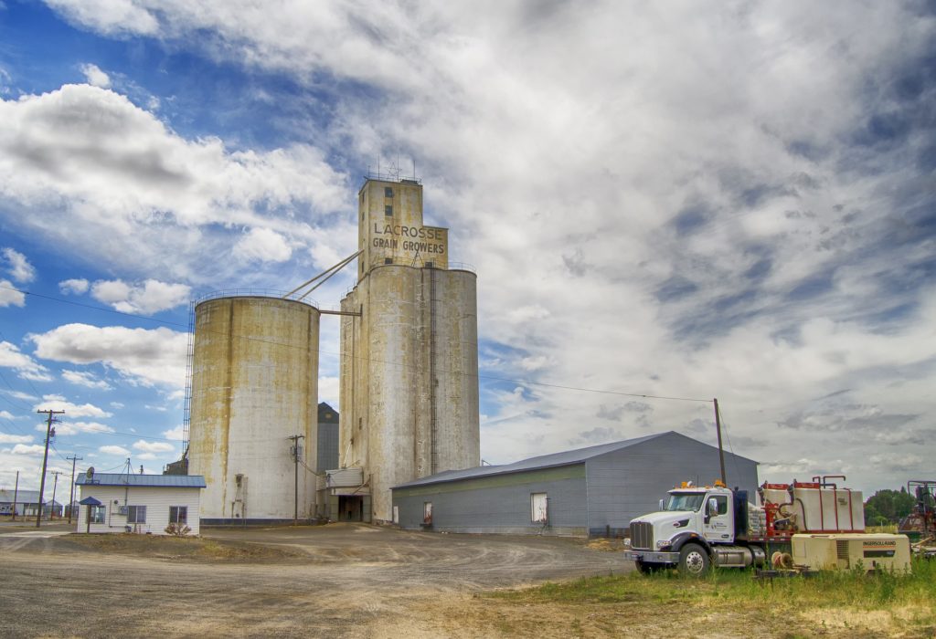 A grain elevator behind a metal shed and a white building.