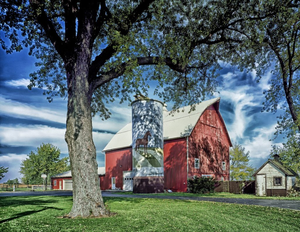 A tall red barn with a grain silo that has a horse painted on its side.
