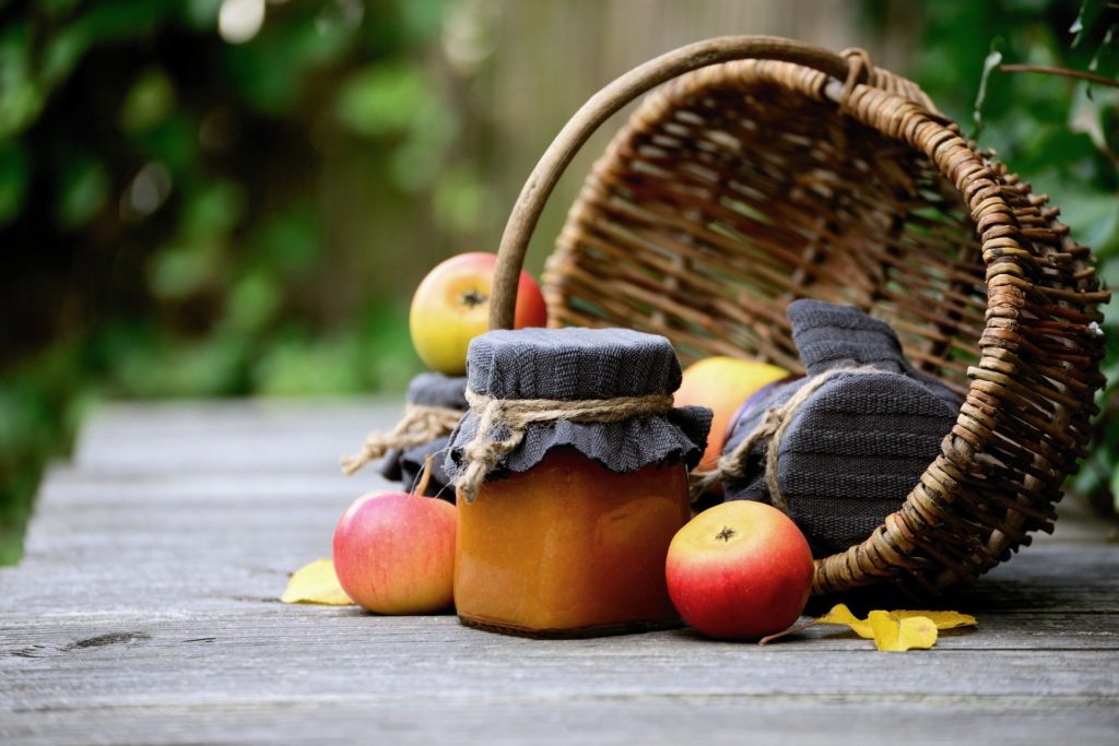 Ripe apples with jars of fresh jam inside a tilted woven basket.