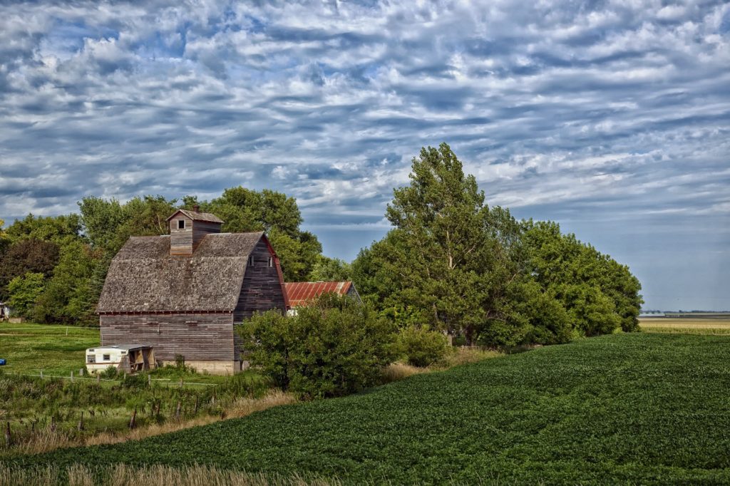 An old barn next to trees and a green field under a cloudy sky.